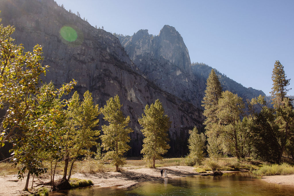 A bride and groom hold hands and look back at the camera while standing on a paved path, with mountains and trees in the background  at Glacier Point Amphitheater