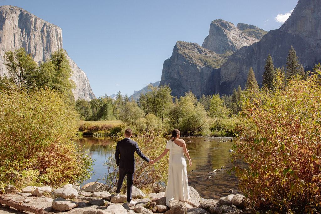 A bride and groom hold hands and look back at the camera while standing on a paved path, with mountains and trees in the background  at Glacier Point Amphitheater