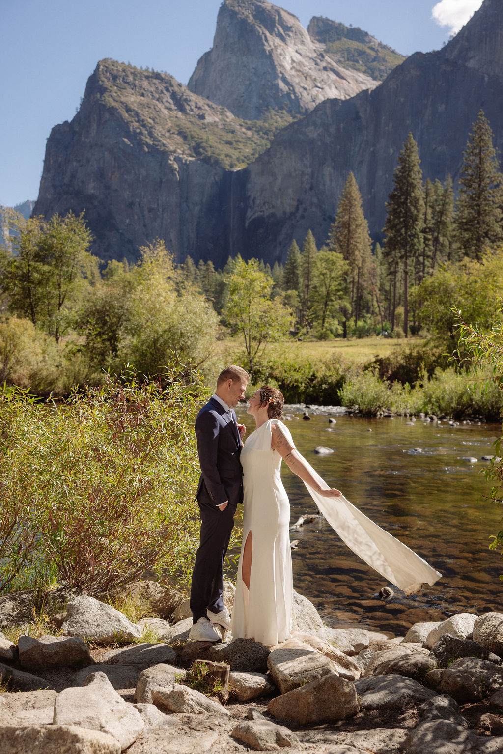 A bride and groom hold hands and look back at the camera while standing on a paved path, with mountains and trees in the background  at Glacier Point Amphitheater