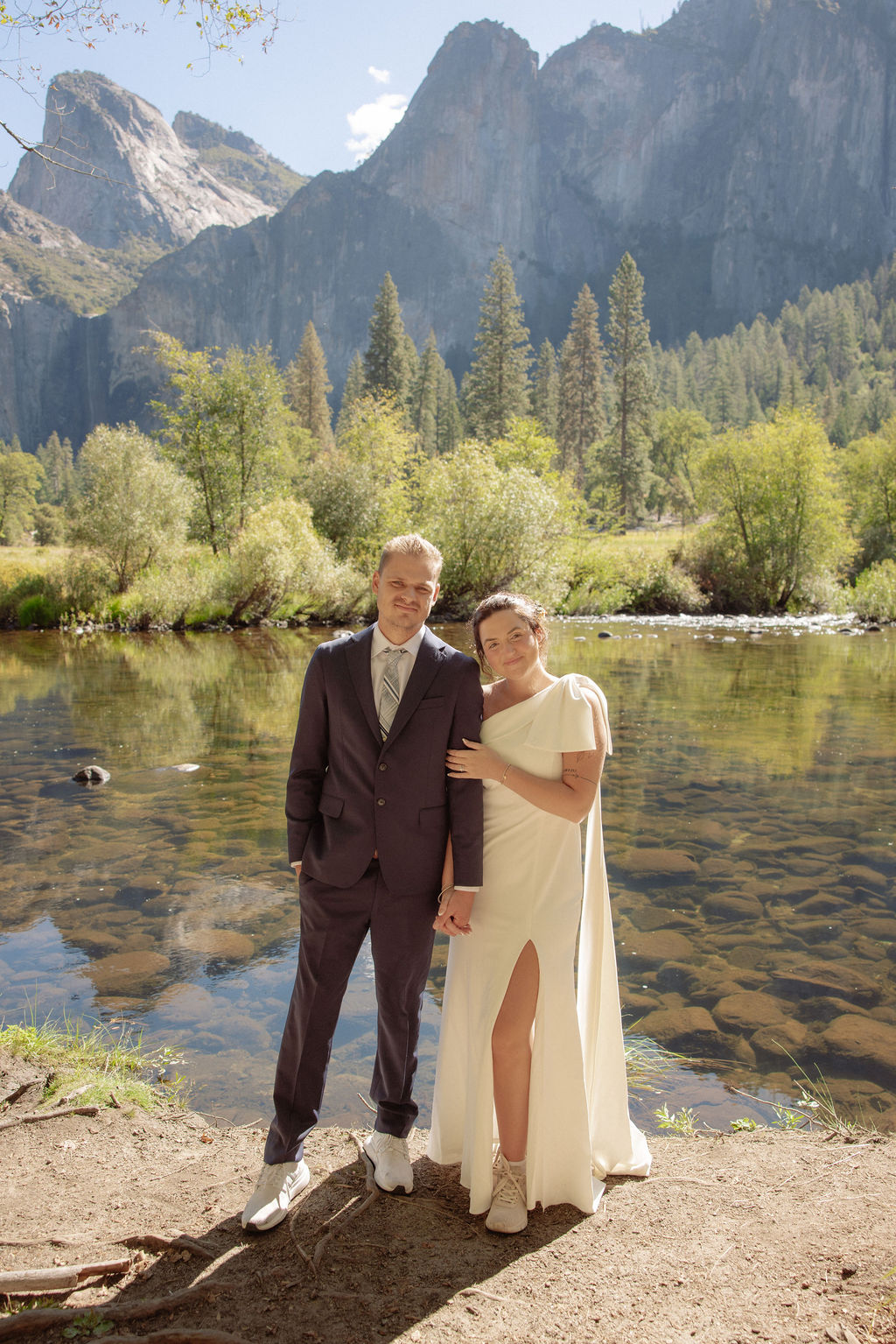 A bride and groom hold hands and look back at the camera while standing on a paved path, with mountains and trees in the background  at Glacier Point Amphitheater