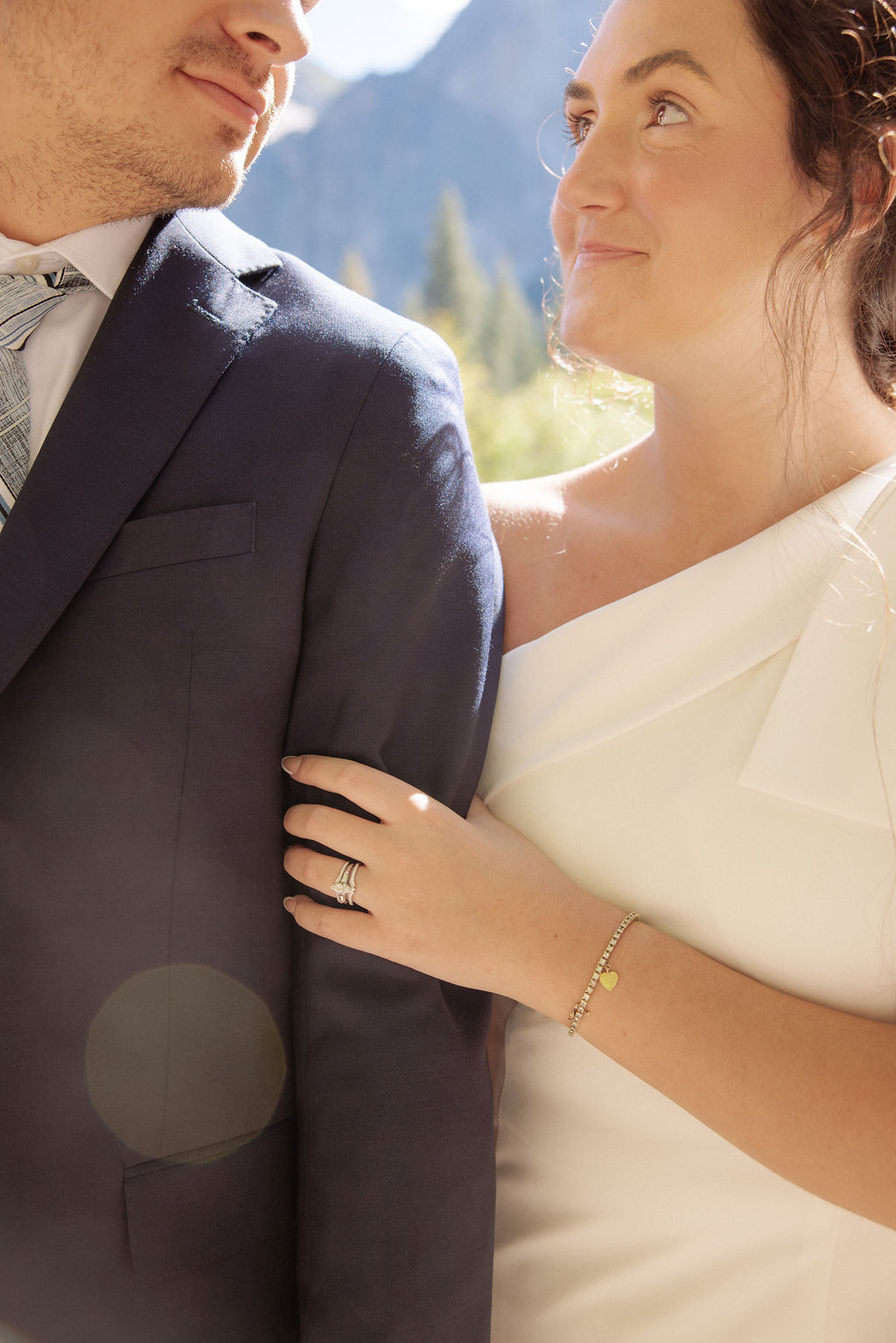 A bride and groom hold hands and look back at the camera while standing on a paved path, with mountains and trees in the background  at Glacier Point Amphitheater