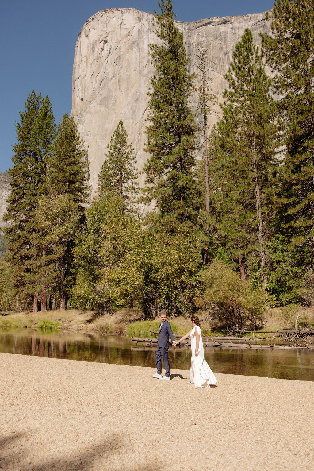 A bride and groom hold hands and look back at the camera while standing on a paved path, with mountains and trees in the background  at Glacier Point Amphitheater