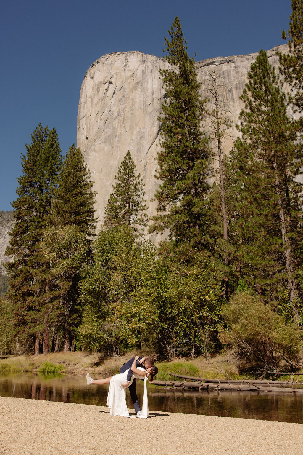 A bride and groom hold hands and look back at the camera while standing on a paved path, with mountains and trees in the background  at Glacier Point Amphitheater