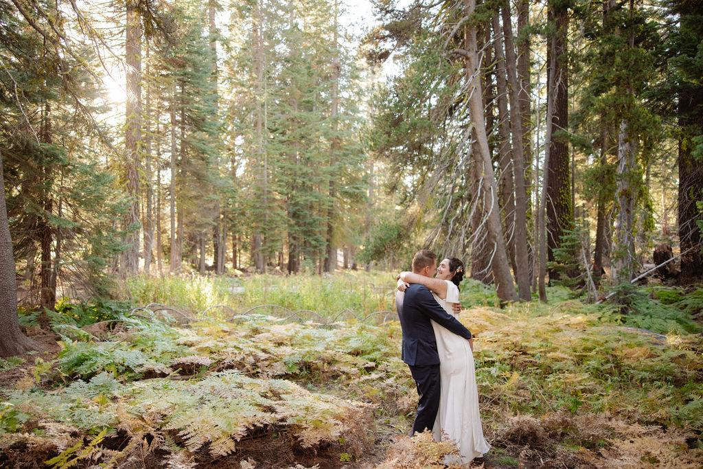 A couple wearing backpacks walks hand-in-hand down a forest trail with sunlight streaming through tall trees.