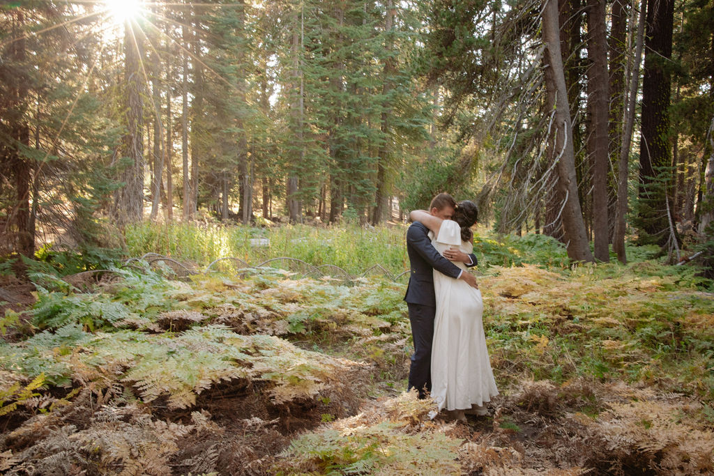 A couple wearing backpacks walks hand-in-hand down a forest trail with sunlight streaming through tall trees.