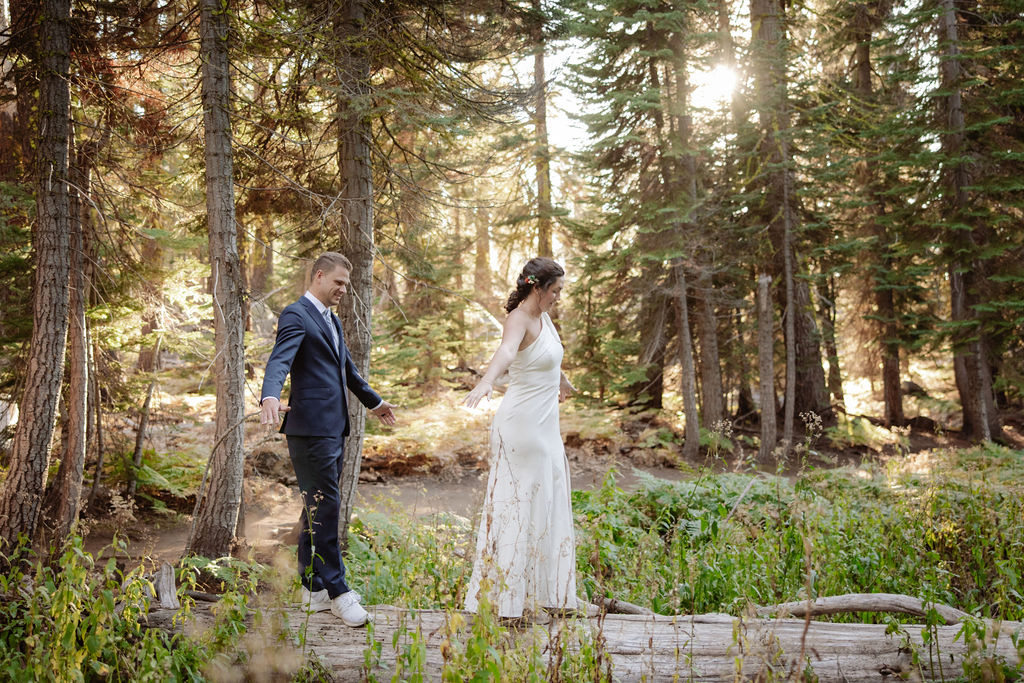 A couple wearing backpacks walks hand-in-hand down a forest trail with sunlight streaming through tall trees.