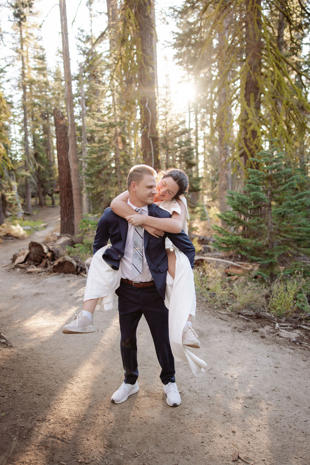 A couple wearing backpacks walks hand-in-hand down a forest trail with sunlight streaming through tall trees.