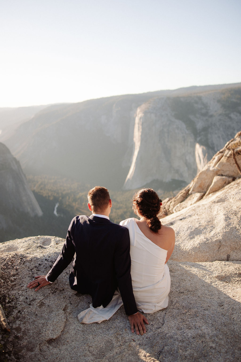 A couple in formal attire sits on a rocky ledge, overlooking a vast mountain landscape under clear skies.