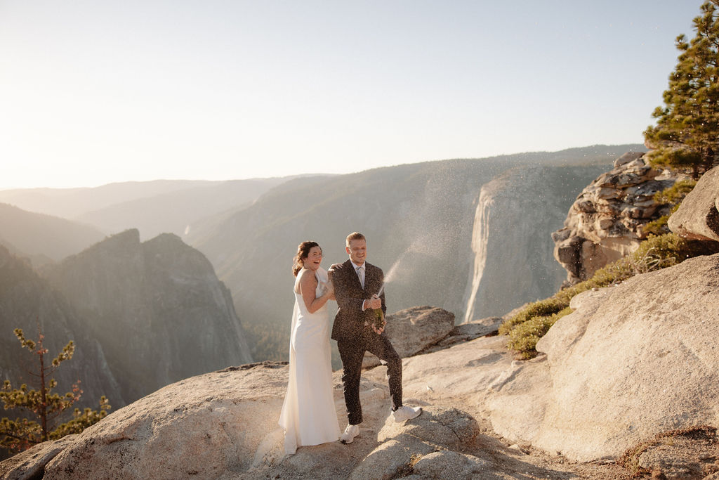 A couple in formal attire sits on a rocky ledge, overlooking a vast mountain landscape under clear skies.