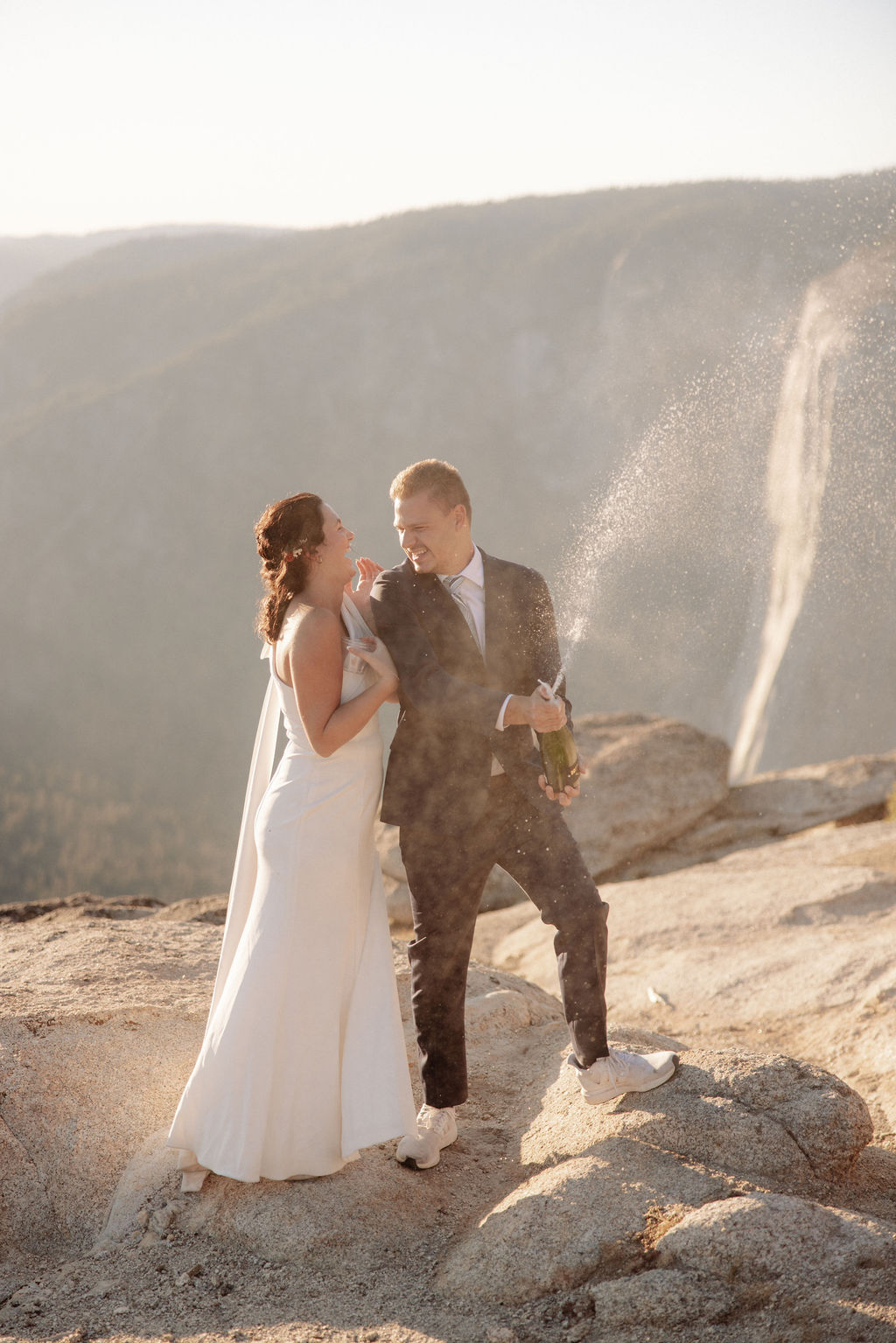 A couple in formal attire sits on a rocky ledge, overlooking a vast mountain landscape under clear skies.
