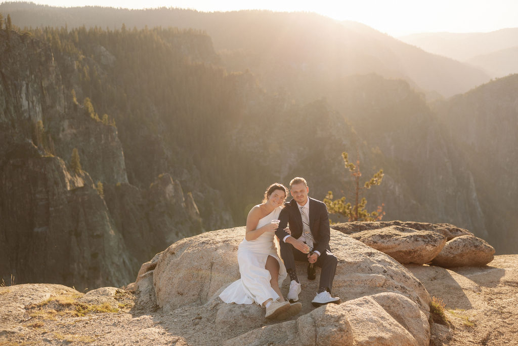 A couple in formal attire sits on a rocky ledge, overlooking a vast mountain landscape under clear skies.