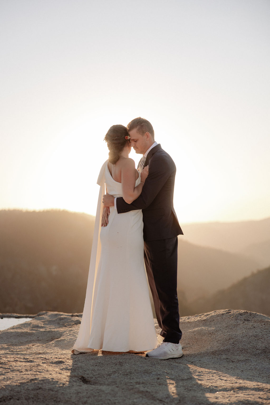 A couple in formal attire sits on a rocky ledge, overlooking a vast mountain landscape under clear skies.