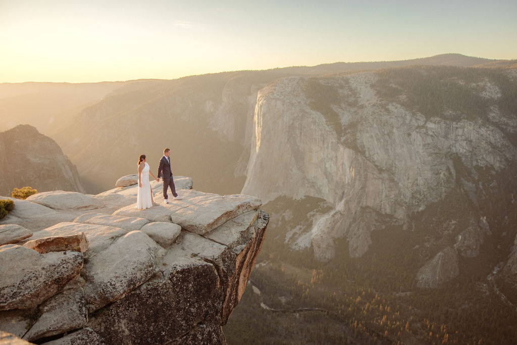 A couple in formal attire sits on a rocky ledge, overlooking a vast mountain landscape under clear skies.