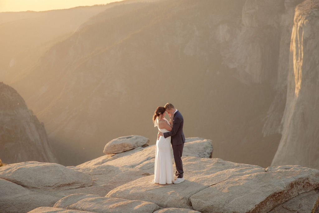 A couple in formal attire sits on a rocky ledge, overlooking a vast mountain landscape under clear skies.