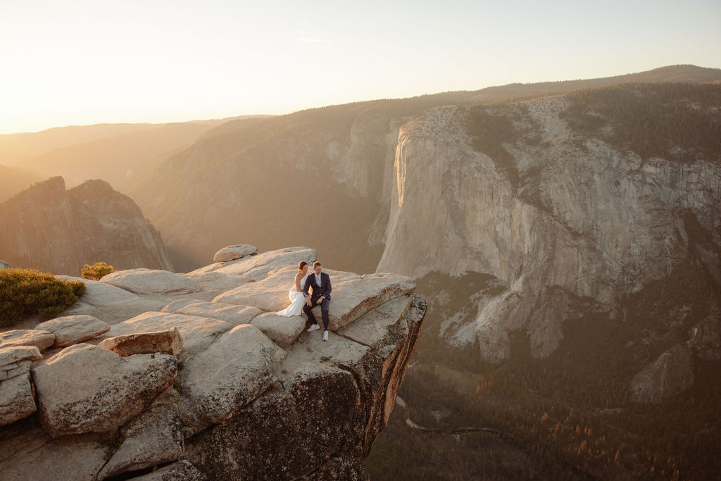 A couple in formal attire sits on a rocky ledge, overlooking a vast mountain landscape under clear skies.
