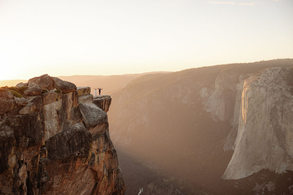 Two people stand near the edge of a high cliff at sunset, overlooking a vast canyon with distant cliffs and soft sunlight.