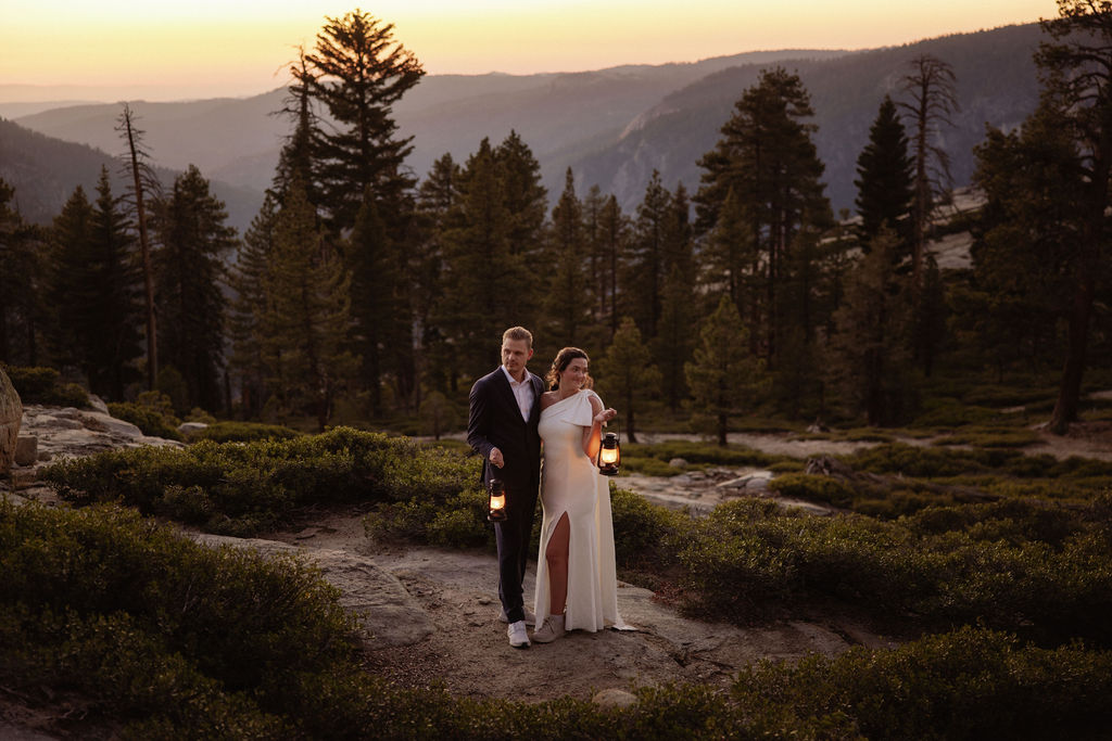 A couple dressed formally stands close together outdoors at sunset, each holding a lit lantern, with trees and hills in the background.