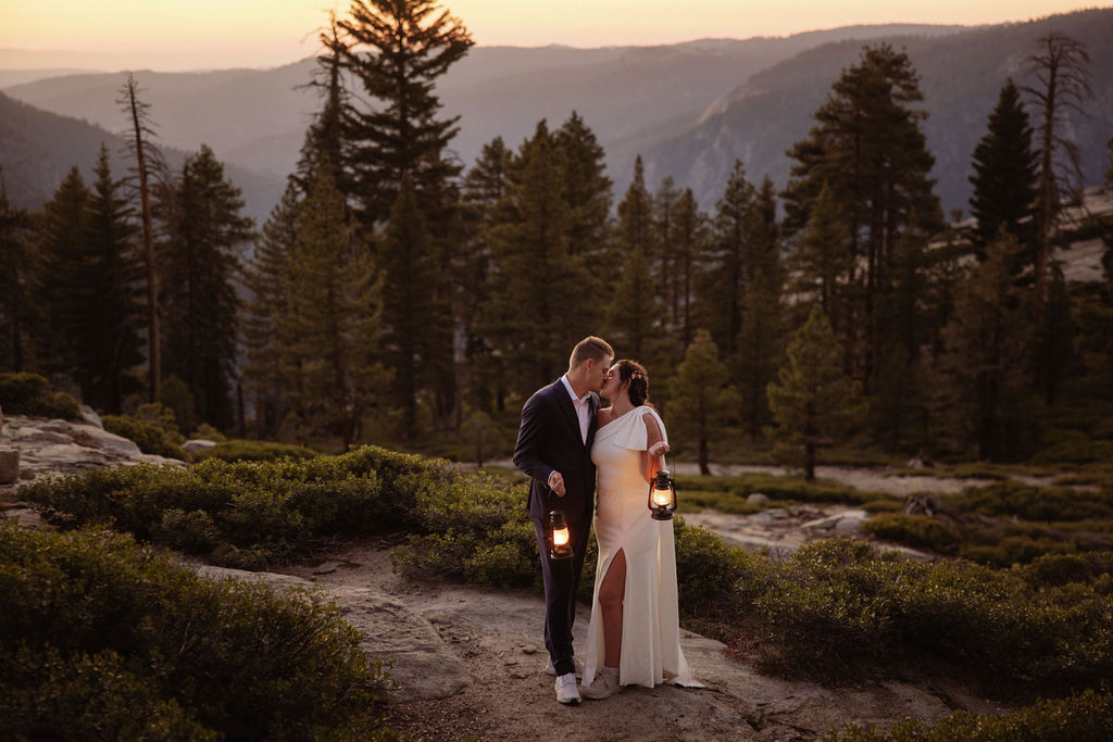 A couple dressed formally stands close together outdoors at sunset, each holding a lit lantern, with trees and hills in the background.