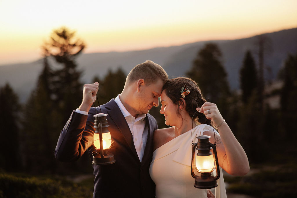 A couple dressed formally stands close together outdoors at sunset, each holding a lit lantern, with trees and hills in the background.