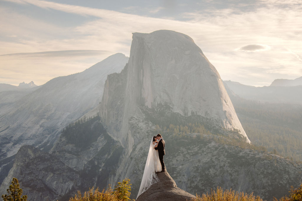 A bride and groom stand on a rocky ledge with a mountain and cloudy sky in the background. The bride holds a bouquet and wears a white dress; the groom wears a black suit.