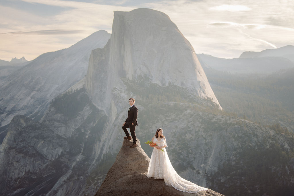 A bride and groom stand on a rocky ledge with a mountain and cloudy sky in the background. The bride holds a bouquet and wears a white dress; the groom wears a black suit.