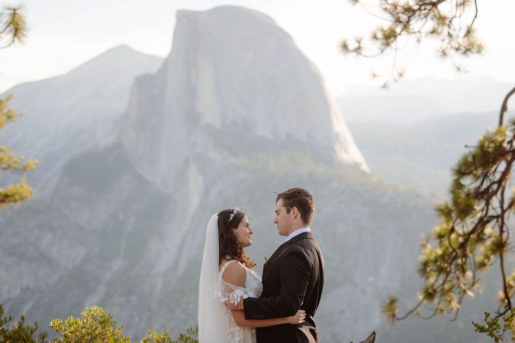 A bride and groom stand on a rocky ledge with a mountain and cloudy sky in the background. The bride holds a bouquet and wears a white dress; the groom wears a black suit.