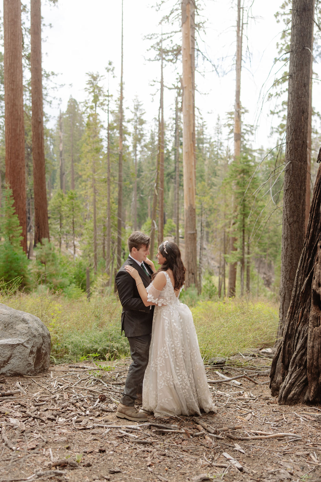 A bride and groom stand together in a forest, looking at each other, surrounded by tall trees and natural greenery for an elopement in Yosemite in September