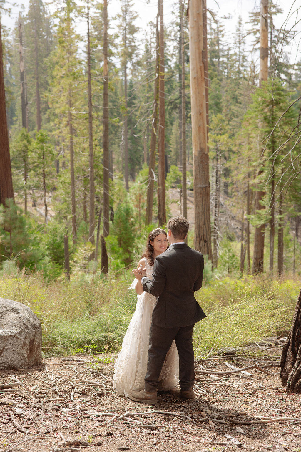 A bride and groom stand together in a forest, looking at each other, surrounded by tall trees and natural greenery for an elopement in Yosemite in September
