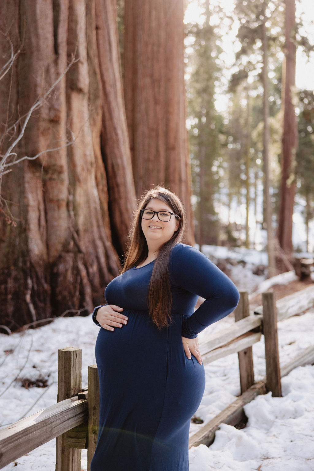 A couple stands close together outdoors near a wooden fence, with tall trees and sunlight in the background; the woman is wearing a long red dress.
