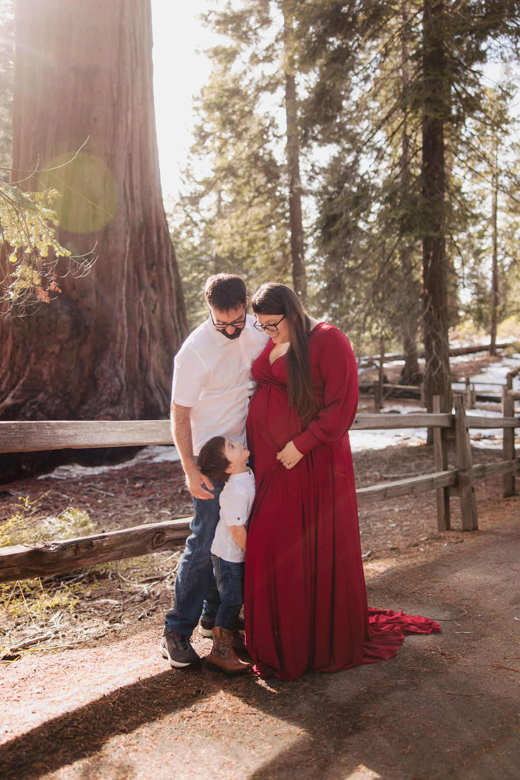 A couple stands close together outdoors near a wooden fence, with tall trees and sunlight in the background; the woman is wearing a long red dress.