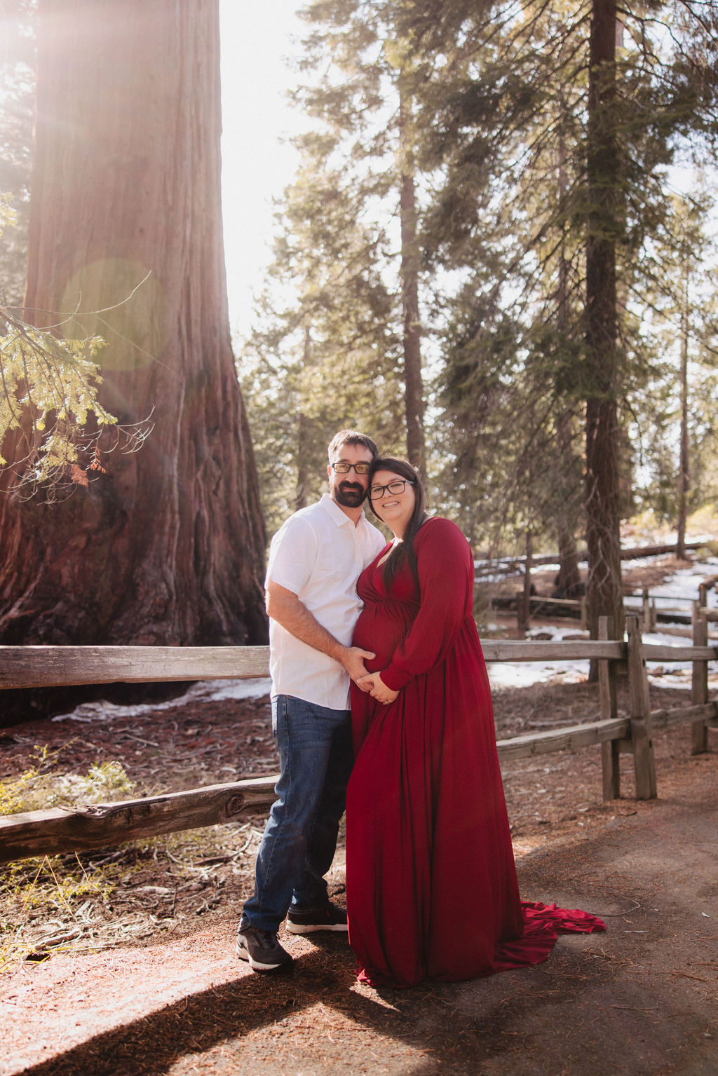 A couple stands close together outdoors near a wooden fence, with tall trees and sunlight in the background; the woman is wearing a long red dress.
