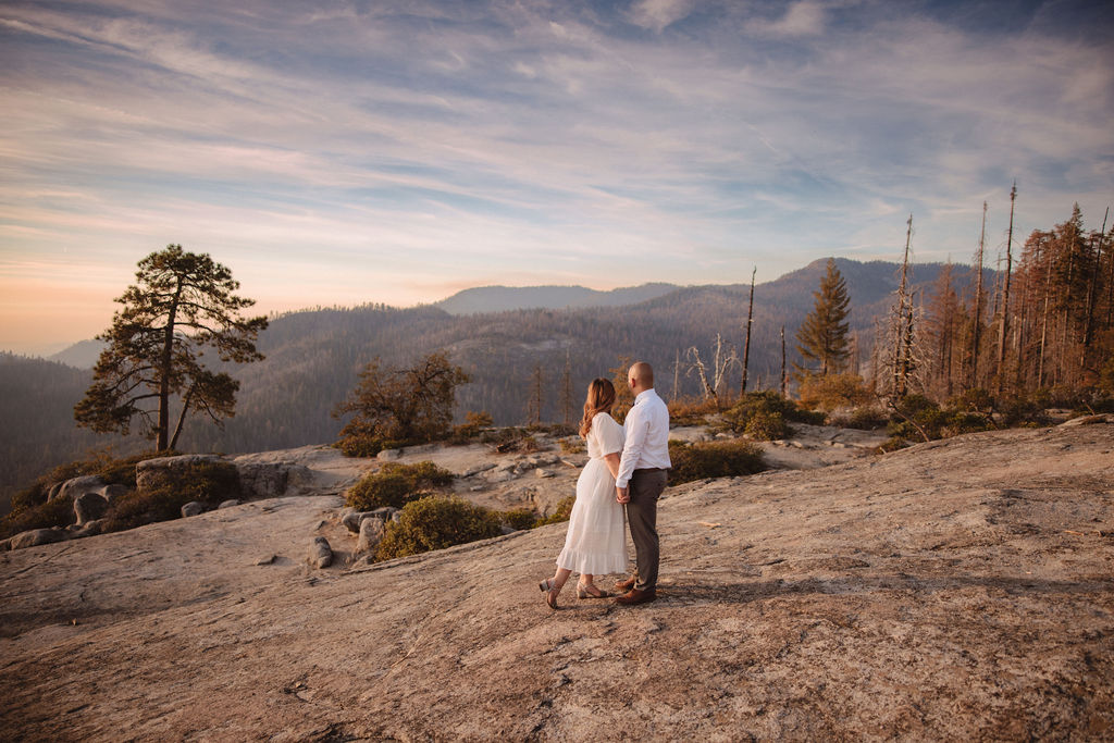 A couple stands close together on a rocky landscape with mountains and trees in the background under a partly cloudy sky.