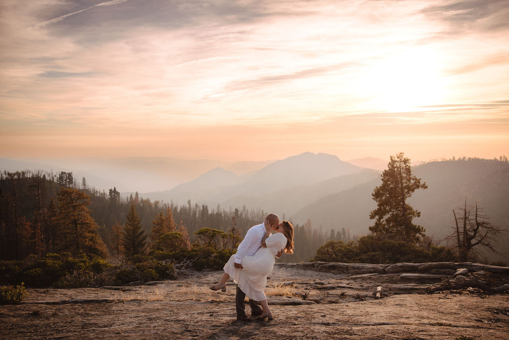 A couple stands close together on a rocky landscape with mountains and trees in the background under a partly cloudy sky.